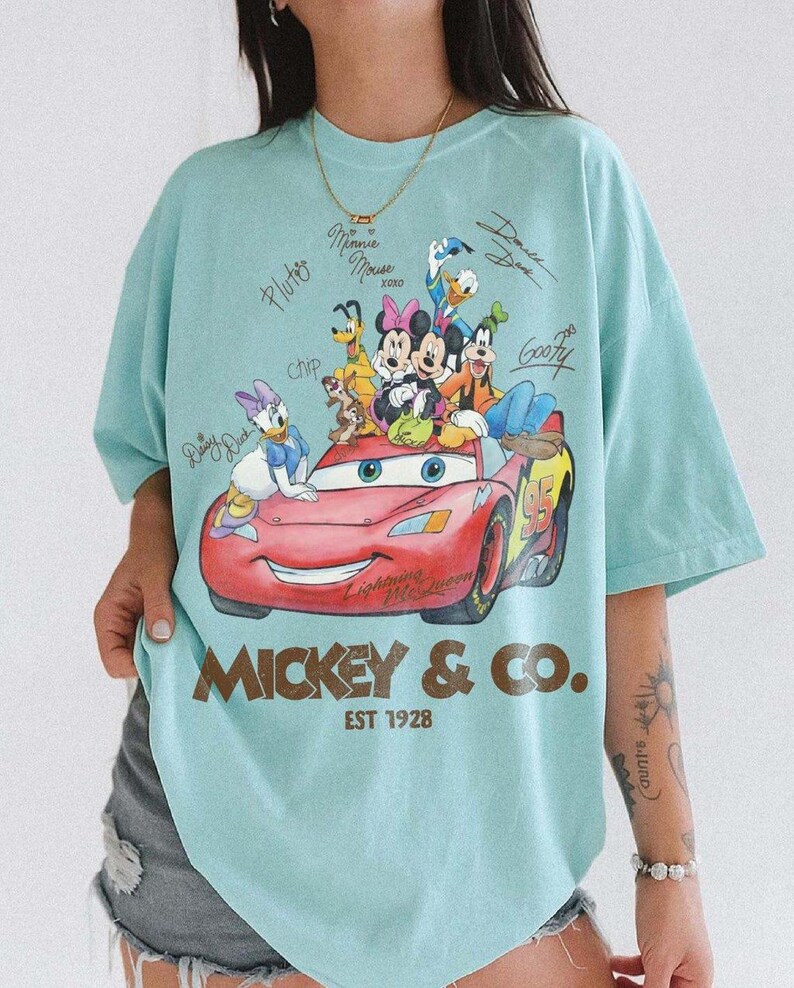 May include: A light blue t-shirt featuring a graphic of Disney characters, including Mickey Mouse, Minnie Mouse, and Donald Duck, on a red race car. The shirt has the text "Mickey & Co. EST 1928".