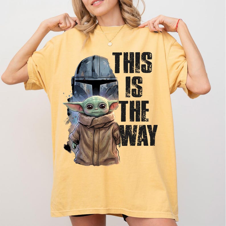 May include: Mustard yellow t-shirt with a graphic of a Mandalorian helmet and Grogu, with the text "THIS IS THE WAY" in black. The design includes a detailed illustration of Grogu in a brown coat. The shirt is a comfortable, casual style.