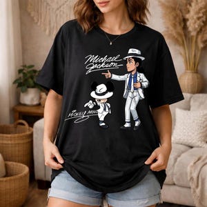 K&ouml;nnte beinhalten: Schwarzes T-Shirt mit einer Cartoon-Illustration von Michael Jackson in einem wei&szlig;en Anzug und Hut, der auf eine Mickey Mouse-Figur zeigt, die ebenfalls in einem &auml;hnlichen Outfit gekleidet ist. Die W&ouml;rter "Michael Jackson" und "Mickey Mouse" sind kursiv geschrieben.