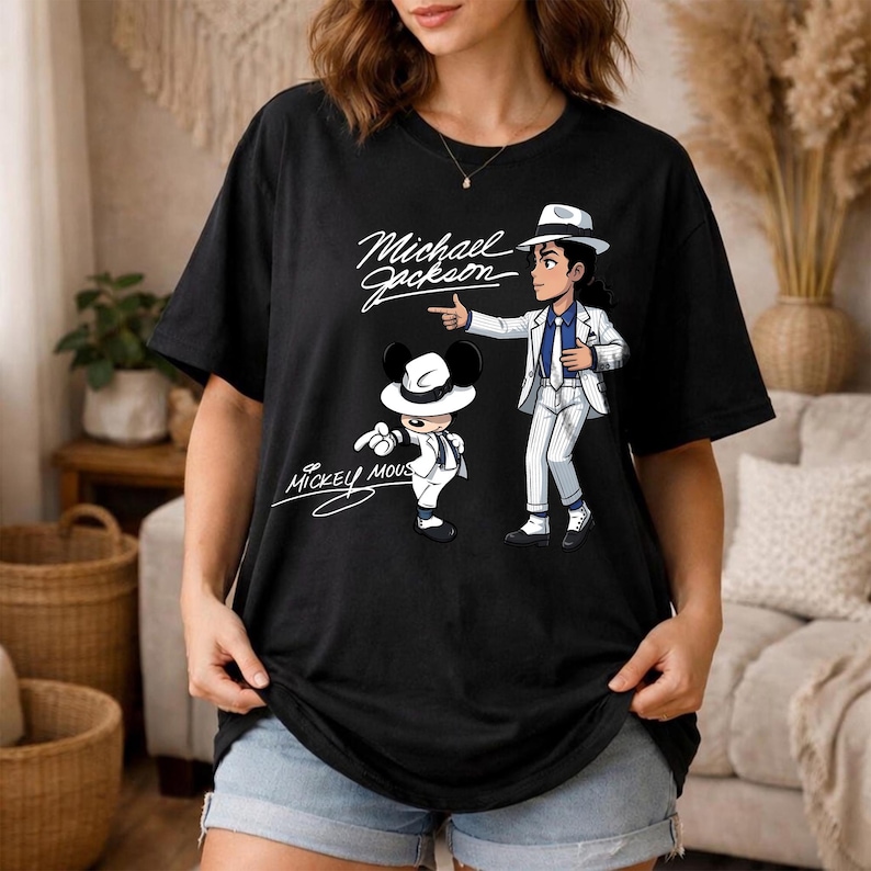 K&ouml;nnte beinhalten: Schwarzes T-Shirt mit einer Cartoon-Illustration von Michael Jackson in einem wei&szlig;en Anzug und Hut, der auf eine Mickey Mouse-Figur zeigt, die ebenfalls in einem &auml;hnlichen Outfit gekleidet ist. Die W&ouml;rter "Michael Jackson" und "Mickey Mouse" sind kursiv geschrieben.
