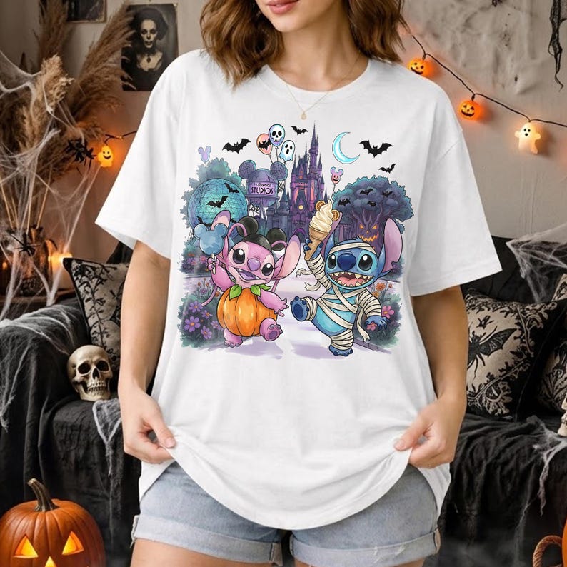 May include: A white t-shirt featuring a graphic of two blue and pink alien characters in Halloween costumes. One character wears a pumpkin suit, the other is wrapped like a mummy. The background shows a castle, bats, and balloons.