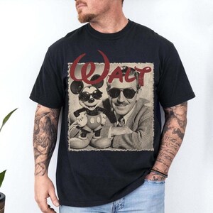 May include: Black t-shirt featuring a sepia-toned image of Walt Disney and Mickey Mouse wearing sunglasses. The word "WALT" is printed in red above the image. The shirt is a classic crew neck style.