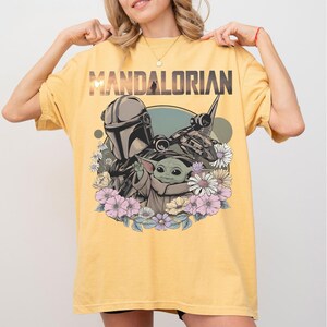 May include: Yellow t-shirt featuring the word "MANDALORIAN" above a graphic of the Mandalorian, Grogu, and a spaceship, surrounded by flowers. The design is in shades of gray, green, and pink.