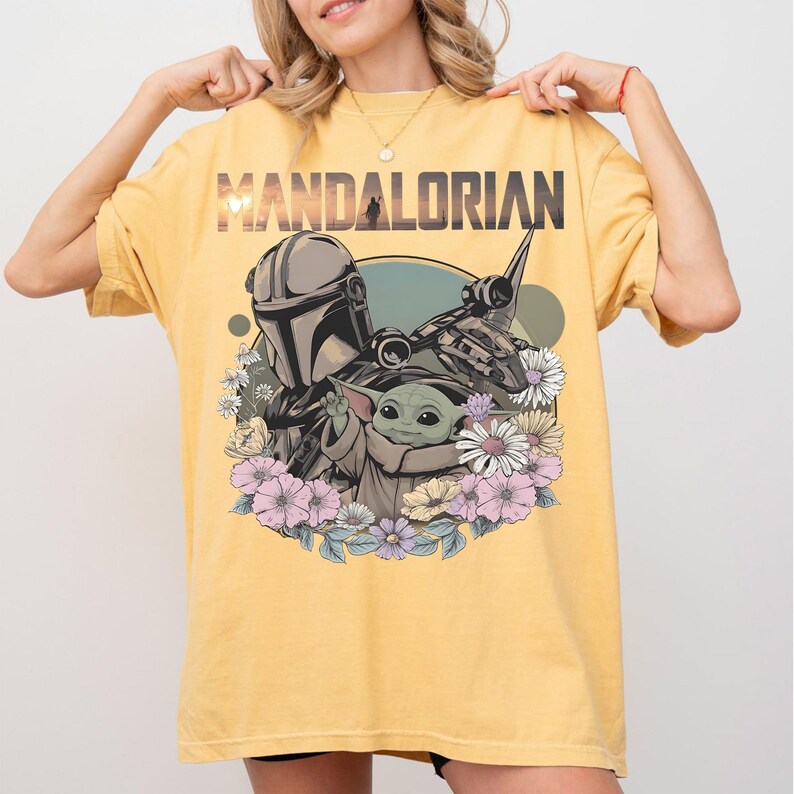 May include: Yellow t-shirt featuring the word "MANDALORIAN" above a graphic of the Mandalorian, Grogu, and a spaceship, surrounded by flowers. The design is in shades of gray, green, and pink.