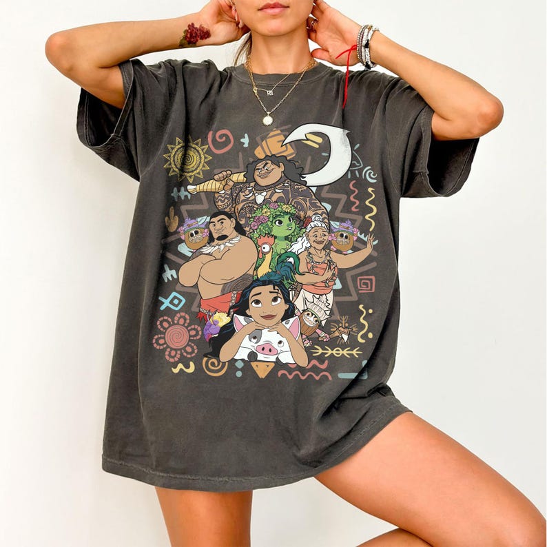 May include: A charcoal gray short-sleeve t-shirt featuring a colorful graphic print of characters from the movie Moana, including Moana, Maui, Pua, Heihei, and Gramma Tala, surrounded by various tribal-inspired symbols and patterns.