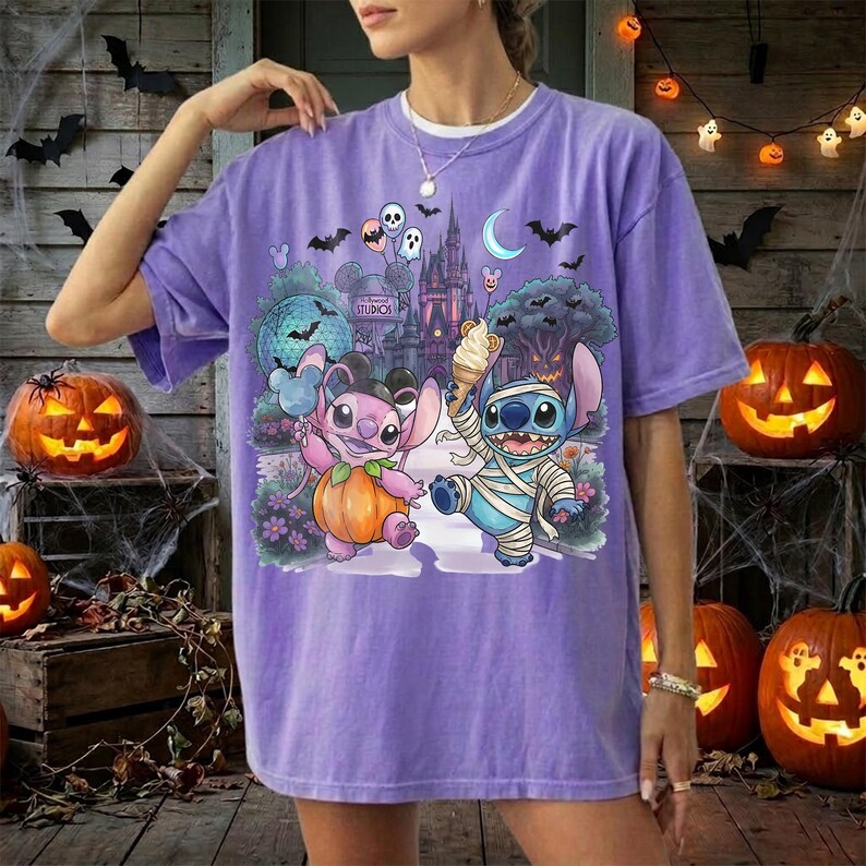 May include: A purple short-sleeve t-shirt featuring a graphic print of two blue alien characters in Halloween costumes. One character holds a pumpkin, while the other is wrapped in bandages like a mummy and holds an ice cream cone. The background shows a castle.