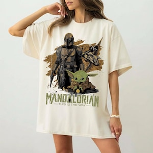 May include: Cream-colored t-shirt featuring a graphic of The Mandalorian, Grogu, and a spaceship. The design includes the text "MANDALORIAN - THIS IS THE WAY" in green and brown tones. The shirt is a casual, oversized fit.
