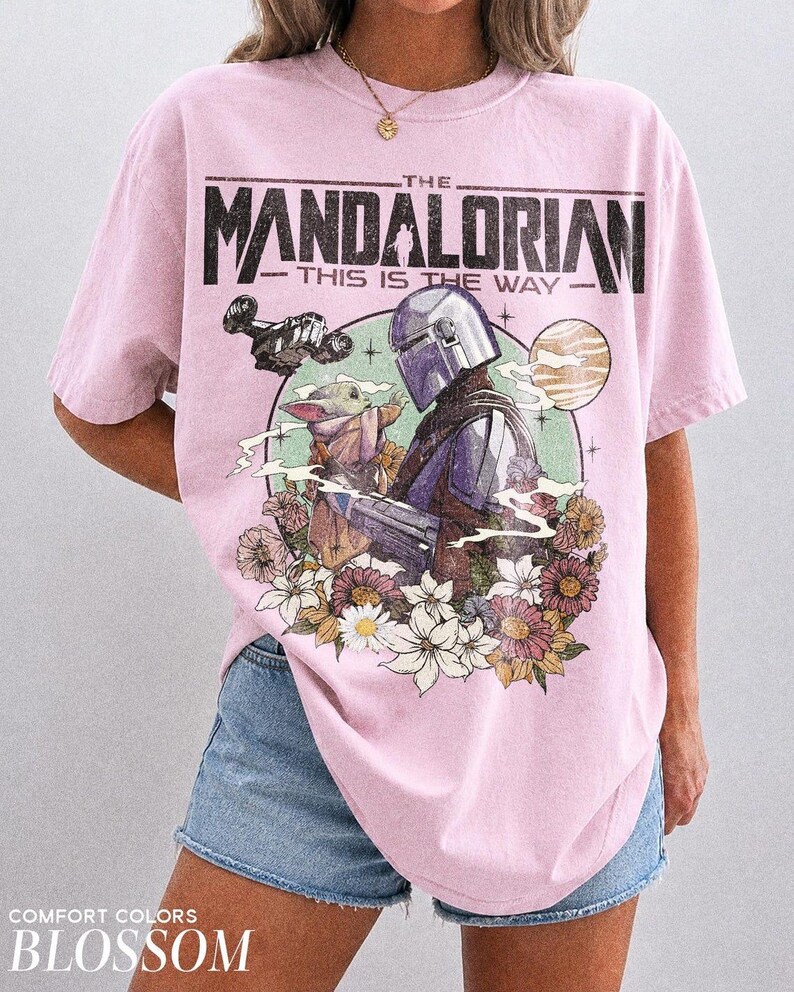 May include: Pale pink t-shirt with a graphic of The Mandalorian and Grogu, framed by flowers. The text "THE MANDALORIAN - THIS IS THE WAY" is printed above the image. The shirt is a Comfort Colors Blossom.