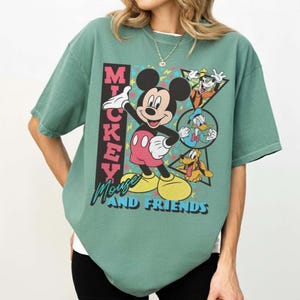 May include: A green t-shirt featuring Mickey Mouse and friends. The graphic includes Mickey Mouse in a red and white outfit, with Donald Duck, Goofy, and Pluto. The text "Mickey Mouse and Friends" is also displayed.