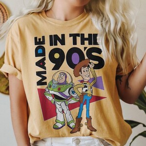 May include: Mustard yellow t-shirt featuring the phrase "MADE IN THE 90'S" in black block letters, with cartoon images of Buzz Lightyear and Woody from Toy Story. The shirt has rolled-up sleeves.