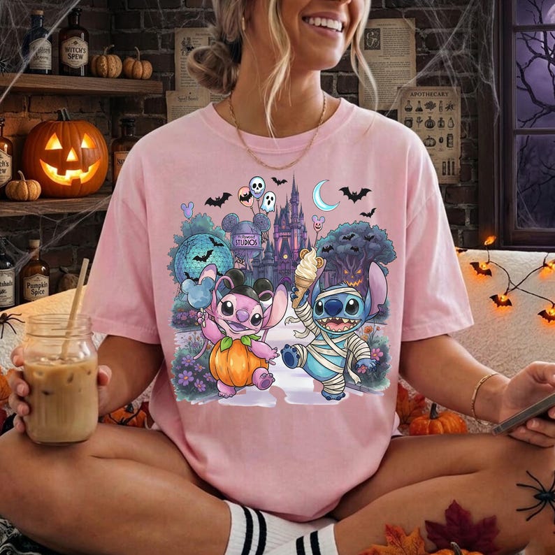 May include: A light pink short-sleeve t-shirt featuring a graphic print of two cartoon characters, one dressed as a pumpkin and one as a mummy, standing in front of a castle with bats, balloons, and a crescent moon in the night sky.