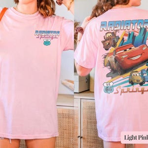 May include: A light pink t-shirt featuring a small graphic on the front chest and a large graphic on the back. The back design displays the text Radiator Springs, a US 66 shield, and illustrations of three cartoon cars in red, brown, and blue.