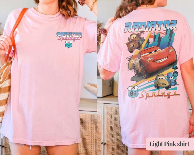 May include: A light pink t-shirt featuring a small graphic on the front chest and a large graphic on the back. The back design displays the text Radiator Springs, a US 66 shield, and illustrations of three cartoon cars in red, brown, and blue.
