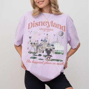Puede incluir: Una camiseta de color lila claro con un gr&aacute;fico de Disneyland. El gr&aacute;fico presenta un castillo, Mickey Mouse, una noria y el texto "Disneyland California Est. 1955" y "The happiest place on earth."