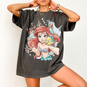Op de afbeelding: Een antracietgrijs T-shirt met korte mouwen en een kleurrijke grafische print van een zeemeermin met rood haar, een gele en blauwe vis, een kleine rode krab en een witte meeuw, omringd door koraal en bubbels in een onderwatersc&egrave;ne.