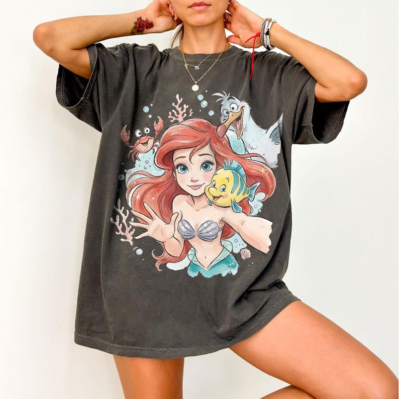 Op de afbeelding: Een antracietgrijs T-shirt met korte mouwen en een kleurrijke grafische print van een zeemeermin met rood haar, een gele en blauwe vis, een kleine rode krab en een witte meeuw, omringd door koraal en bubbels in een onderwatersc&egrave;ne.