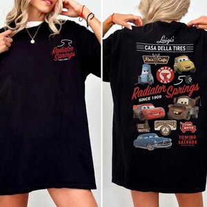 May include: A black t-shirt featuring a small Radiator Springs logo on the front chest. The back displays a large graphic with various characters and logos, including Luigi's Casa Della Tires, Flo's Cafe, and the text Radiator Springs Since 1909.