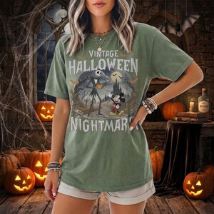K&ouml;nnte beinhalten: Ein salbeigr&uuml;nes Kurzarm-T-Shirt mit einem Grafikdruck von Jack Skellington und Mickey Mouse in einer gruseligen Landschaft. Der Text lautet VINTAGE HALLOWEEN NIGHTMARE. Das Design zeigt einen Vollmond, Flederm&auml;use und eine dunkle Burgsilhouette.