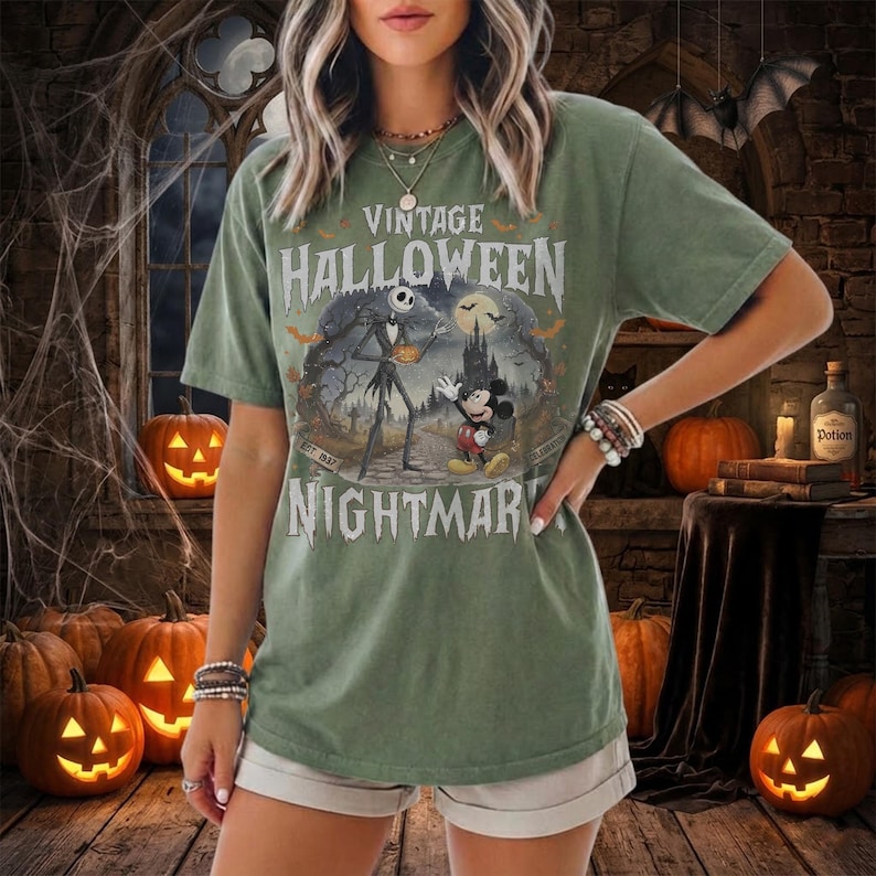 K&ouml;nnte beinhalten: Ein salbeigr&uuml;nes Kurzarm-T-Shirt mit einem Grafikdruck von Jack Skellington und Mickey Mouse in einer gruseligen Landschaft. Der Text lautet VINTAGE HALLOWEEN NIGHTMARE. Das Design zeigt einen Vollmond, Flederm&auml;use und eine dunkle Burgsilhouette.