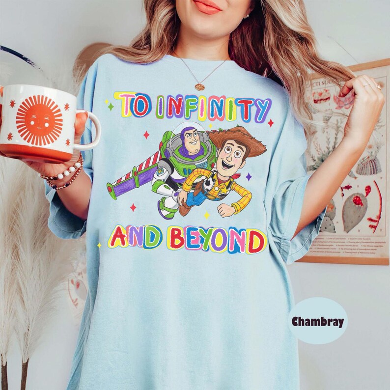 May include: Light blue t-shirt featuring the phrase "TO INFINITY AND BEYOND" in colorful letters, with illustrations of Buzz Lightyear and Woody. The shirt has a relaxed fit and is made of a soft material. The word "Chambray" is printed on the bottom right.