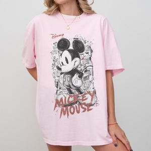 Puede incluir: Camiseta rosa claro con un dise&ntilde;o en blanco y negro de Mickey Mouse. El dise&ntilde;o incluye la palabra "Disney" y "Mickey Mouse" en un estilo esbozado. La camiseta tiene mangas cortas.
