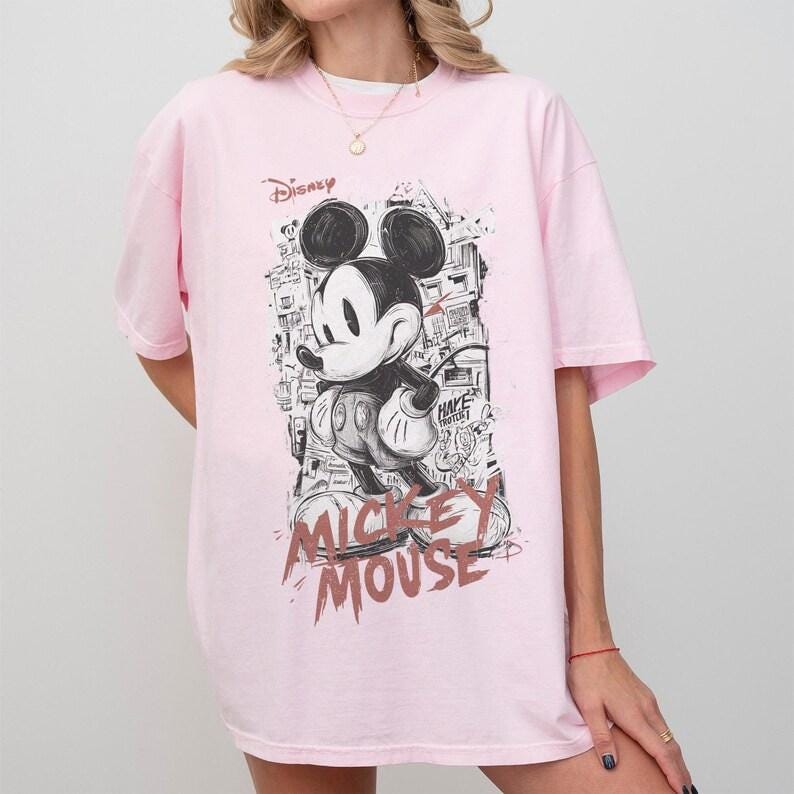 Puede incluir: Camiseta rosa claro con un dise&ntilde;o en blanco y negro de Mickey Mouse. El dise&ntilde;o incluye la palabra "Disney" y "Mickey Mouse" en un estilo esbozado. La camiseta tiene mangas cortas.