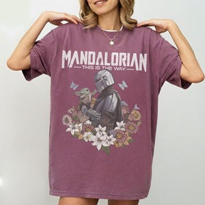 K&ouml;nnte beinhalten: Ein weinrotes T-Shirt mit dem wei&szlig;en Schriftzug "MANDALORIAN - THIS IS THE WAY". Die Grafik zeigt den Mandalorianer und Grogu mit Blumen und Schmetterlingen. Das T-Shirt hat eine lockere Passform.