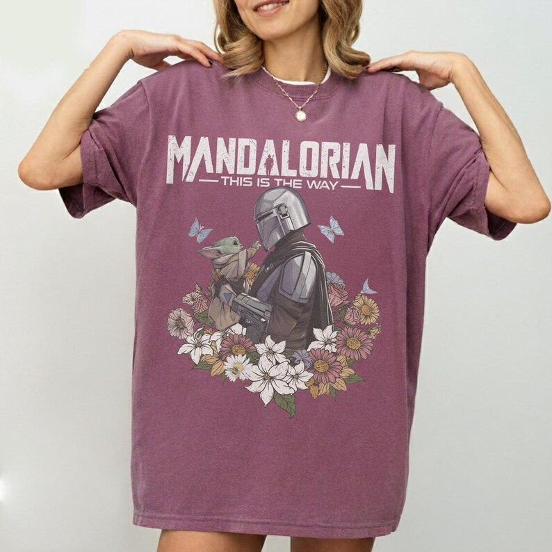 K&ouml;nnte beinhalten: Ein weinrotes T-Shirt mit dem wei&szlig;en Schriftzug "MANDALORIAN - THIS IS THE WAY". Die Grafik zeigt den Mandalorianer und Grogu mit Blumen und Schmetterlingen. Das T-Shirt hat eine lockere Passform.