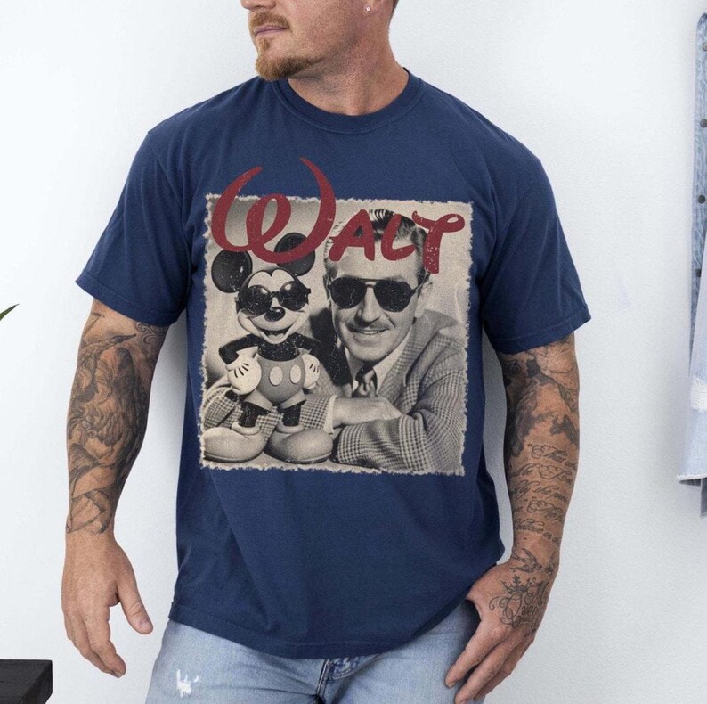 May include: Navy blue t-shirt featuring a vintage-style graphic of Mickey Mouse and Walt Disney wearing sunglasses. The word "WALT" is arched above the image in a burgundy color. The image is a sepia-toned photograph.
