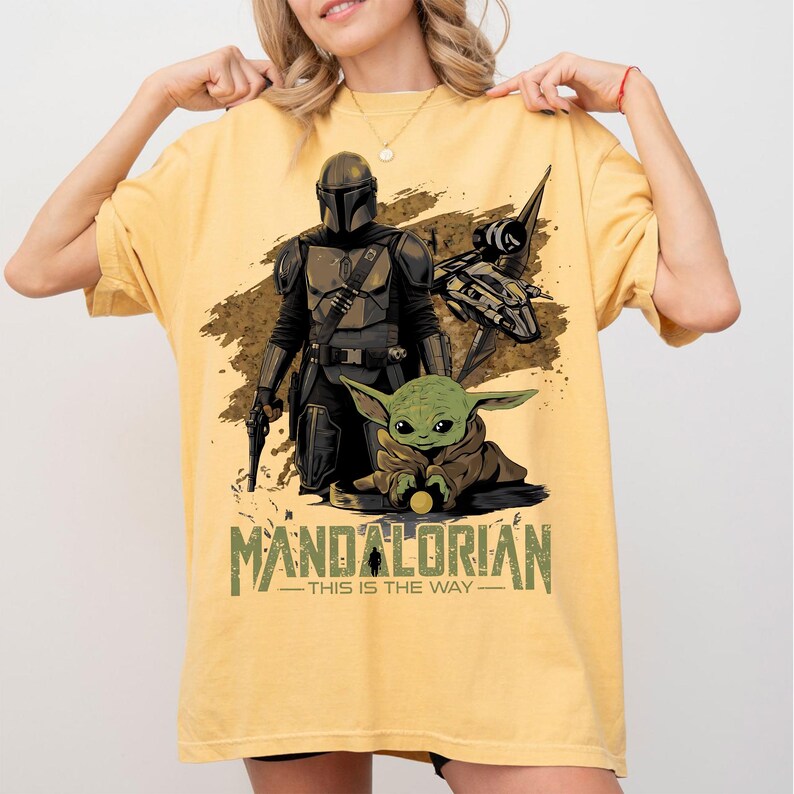 May include: Yellow t-shirt featuring a graphic of The Mandalorian and Grogu, with a spaceship and the text "MANDALORIAN - THIS IS THE WAY" in green and brown. The design includes a detailed illustration of the characters.