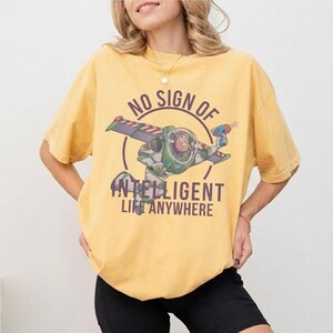 Pu&ograve; includere: T-shirt giallo senape con una grafica di Buzz Lightyear di Toy Story. La grafica include il testo "NO SIGN OF INTELLIGENT LIFE ANYWHERE" in un design circolare. La maglietta ha maniche corte e girocollo.