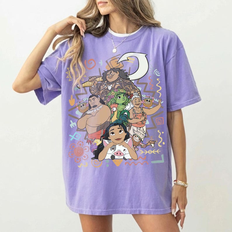 May include: A light purple short-sleeve t-shirt featuring a large graphic print of various animated characters including a large man with a hook, a woman, a small pig, and a rooster, surrounded by colorful geometric shapes and tribal patterns.