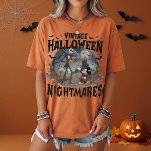 K&ouml;nnte beinhalten: Ein orangefarbenes Kurzarm-T-Shirt mit einem Grafikdruck und dem Text VINTAGE HALLOWEEN NIGHTMARES. Das Design zeigt Jack Skellington und Mickey Mouse in einer gruseligen Landschaft mit Schloss, Flederm&auml;usen und dem Text EST. 1937 CELEBRATION.