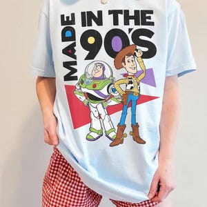 May include: Light blue t-shirt featuring Buzz Lightyear and Woody from Toy Story, with the text "MADE IN THE 90'S". The shirt has a graphic design with red, purple, and white accents. The person is wearing red and white checkered shorts.