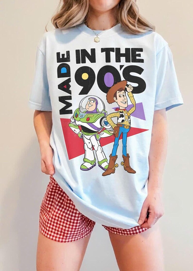 May include: Light blue t-shirt featuring Buzz Lightyear and Woody from Toy Story, with the text "MADE IN THE 90'S". The shirt has a graphic design with red, purple, and white accents. The person is wearing red and white checkered shorts.