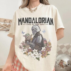 K&ouml;nnte beinhalten: Cremefarbenes T-Shirt mit einer Grafik von The Mandalorian und Grogu, umgeben von Blumen und Schmetterlingen. Der Text "MANDALORIAN - THIS IS THE WAY" ist oben in einer kr&auml;ftigen, verwitterten Schriftart aufgedruckt. Ein stilvolles T-Shirt f&uuml;r Fans der Serie.