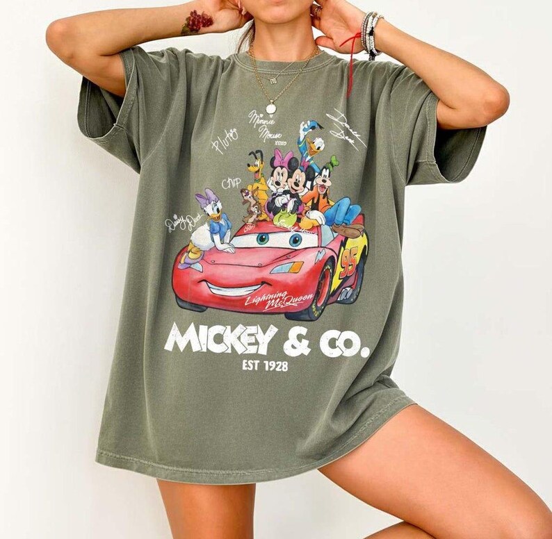 May include: Olive green t-shirt featuring a graphic of Disney characters including Mickey Mouse, Minnie Mouse, Donald Duck, and Lightning McQueen. The text "Mickey & Co. EST 1928" is printed below the graphic.