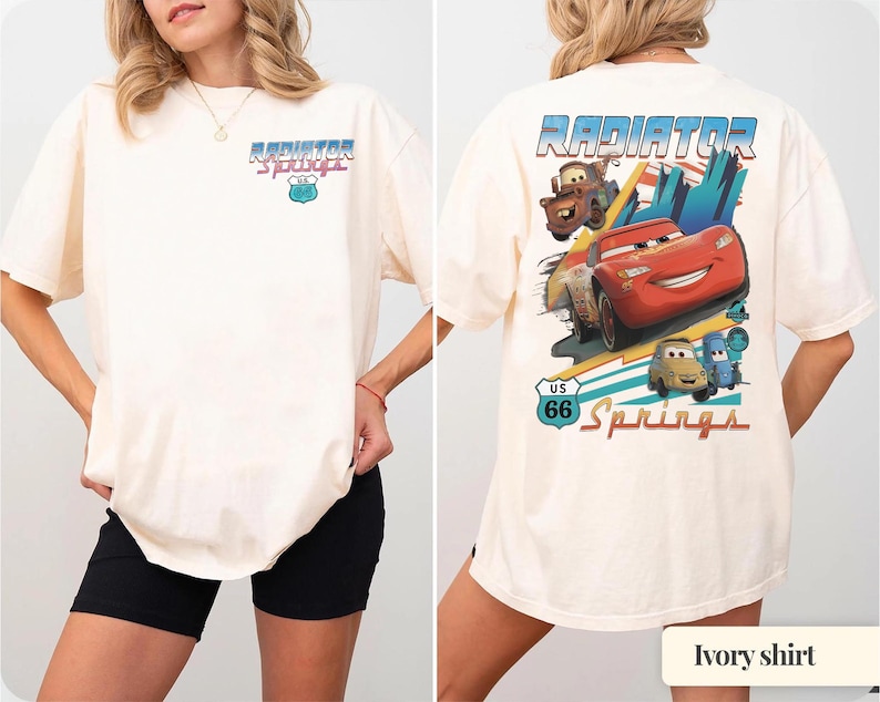 May include: An ivory t-shirt featuring a small Radiator Springs Route 66 graphic on the front chest. The back displays a large, colorful illustration of Lightning McQueen, Mater, and other characters with the text Radiator Springs US 66.