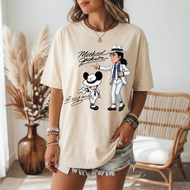 K&ouml;nnte beinhalten: Cremefarbenes T-Shirt mit einer Cartoon-Illustration von Michael Jackson und Mickey Mouse. Das Design enth&auml;lt den Text "Michael Jackson" und "Mickey Mouse" in einer Schriftart. Das Shirt hat kurze &Auml;rmel und eine lockere Passform.