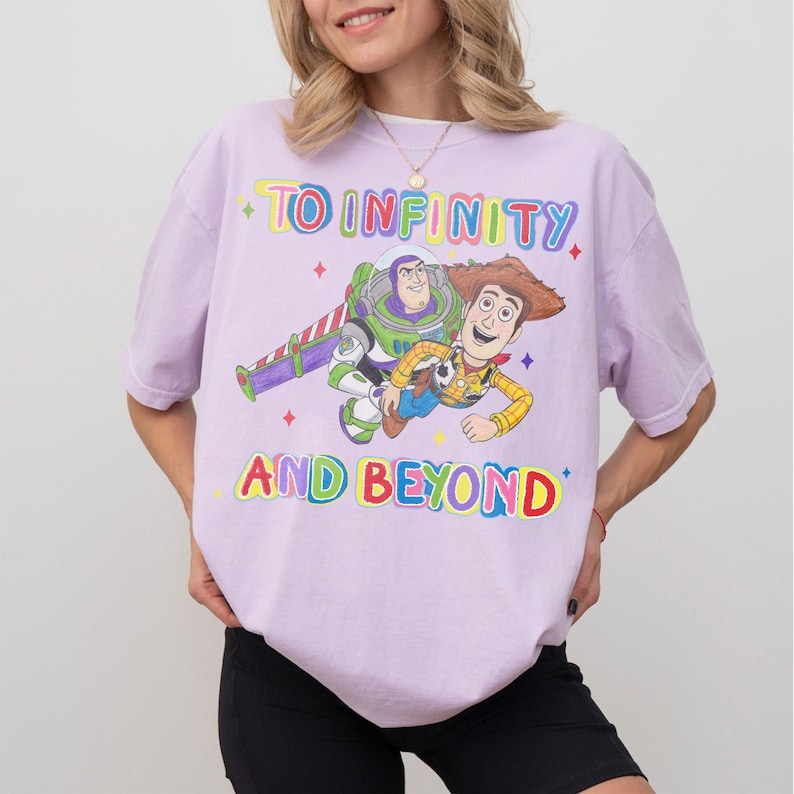 May include: A light purple t-shirt featuring a graphic of Buzz Lightyear and Woody from Toy Story, with the text "TO INFINITY AND BEYOND" in colorful letters. The design includes stars and a rocket. The shirt is a casual wear item.