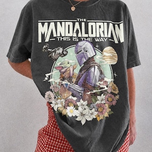 May include: Dark grey t-shirt with a graphic of The Mandalorian and Grogu surrounded by flowers. The text "THE MANDALORIAN - THIS IS THE WAY" is printed across the top. The shirt is labelled "COMFORT COLORS - PEPPER".