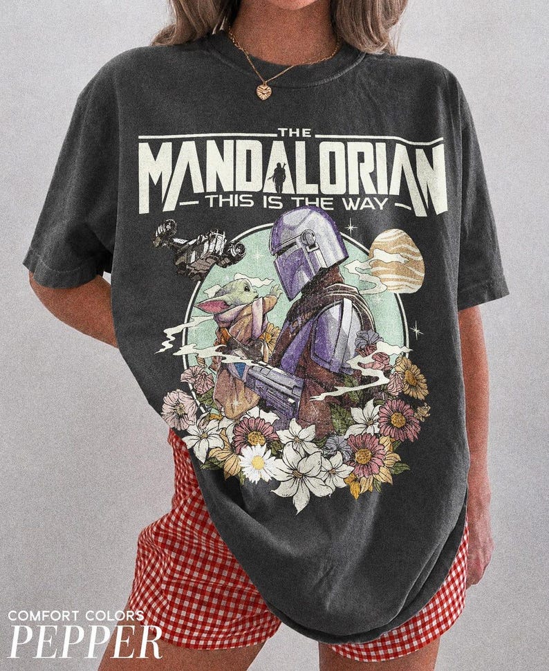 May include: Dark grey t-shirt with a graphic of The Mandalorian and Grogu surrounded by flowers. The text "THE MANDALORIAN - THIS IS THE WAY" is printed across the top. The shirt is labelled "COMFORT COLORS - PEPPER".