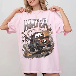 May include: A light pink t-shirt featuring a graphic of Mater from the movie Cars. The design shows Mater in action, kicking up dust and debris. The word "MATER" is printed above the image in a distressed font.