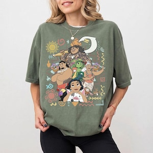 May include: An olive green short-sleeve t-shirt featuring a colorful graphic print of various animated characters, including a central figure holding a large hook, a pig, and tribal-inspired patterns like suns, spirals, and geometric shapes.