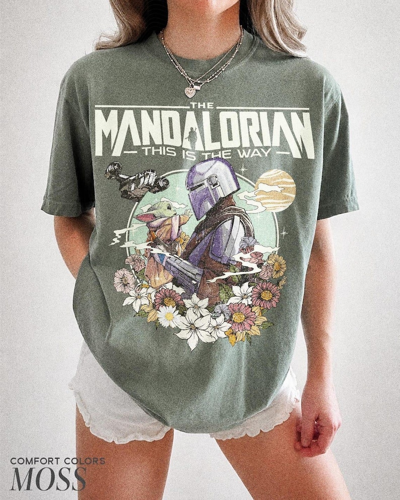 May include: Moss green t-shirt with a graphic of The Mandalorian and Grogu surrounded by flowers. The text "THE MANDALORIAN - THIS IS THE WAY" is printed across the top. The shirt is a Comfort Colors brand.