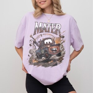 May include: A light purple t-shirt featuring a graphic of Mater from the movie Cars. The word "MATER" is printed above the image of the character. The graphic shows Mater kicking up dirt and debris.