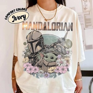 May include: Ivory-colored t-shirt featuring a graphic of The Mandalorian and Grogu surrounded by flowers. The word "MANDALORIAN" is printed across the top. The shirt also has the words "COMFORT COLORS" and "Ivory" in a circular design.