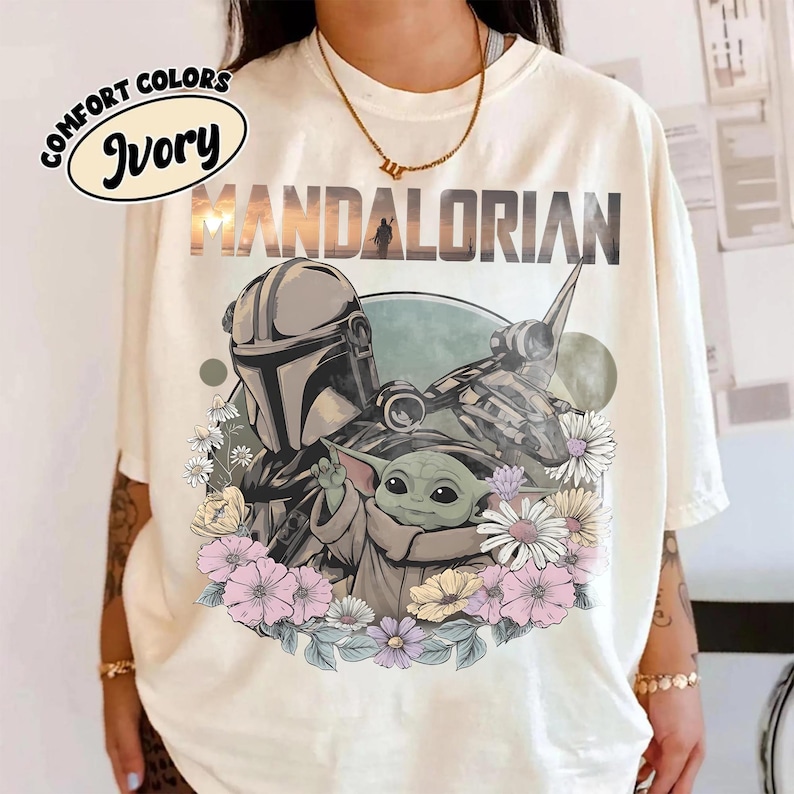 May include: Ivory-colored t-shirt featuring a graphic of The Mandalorian and Grogu surrounded by flowers. The word "MANDALORIAN" is printed across the top. The shirt also has the words "COMFORT COLORS" and "Ivory" in a circular design.