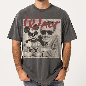 May include: Dark gray t-shirt featuring a black and white image of Walt Disney and Mickey Mouse wearing sunglasses. The word "WALT" is printed in red above the image. The shirt is a vintage style.