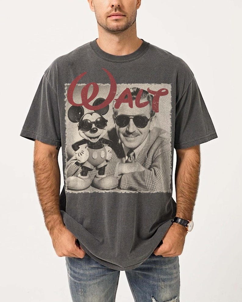 May include: Dark gray t-shirt featuring a black and white image of Walt Disney and Mickey Mouse wearing sunglasses. The word "WALT" is printed in red above the image. The shirt is a vintage style.